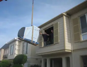 A-large-wrapped-item-is-being-hoisted-out-of-an-upper-window-by-a-crane-while-two-people-assist-from-inside-the-house